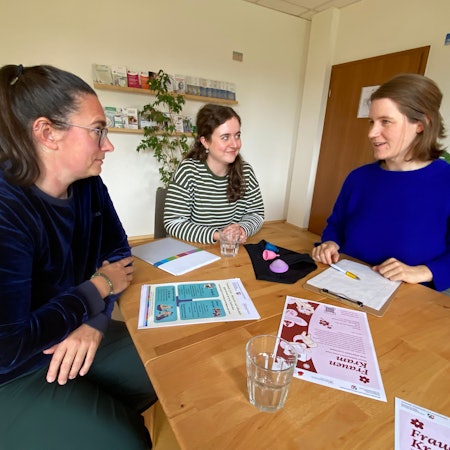 Drei Frauen unterhalten sich. Sie sitzen um einen Tisch, auf dem Unterlagen liegen.