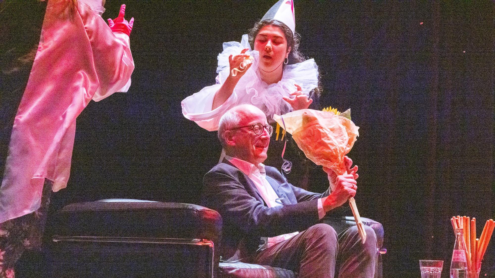 Eine Frau im Clownskostüm bläst eine Luftschlange, ein Mann schaut auf einen Blumenstrauß.