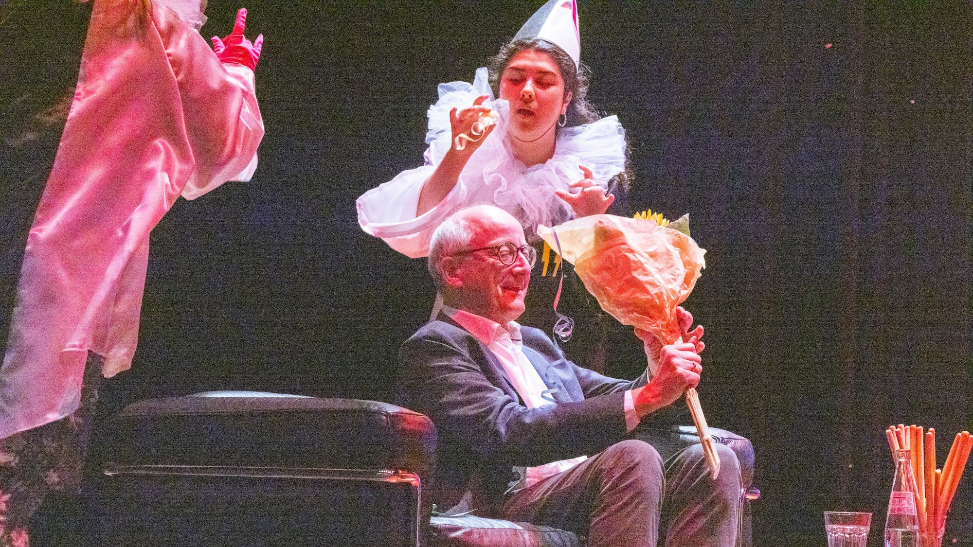 Eine Frau im Clownskostüm bläst eine Luftschlange, ein Mann schaut auf einen Blumenstrauß.