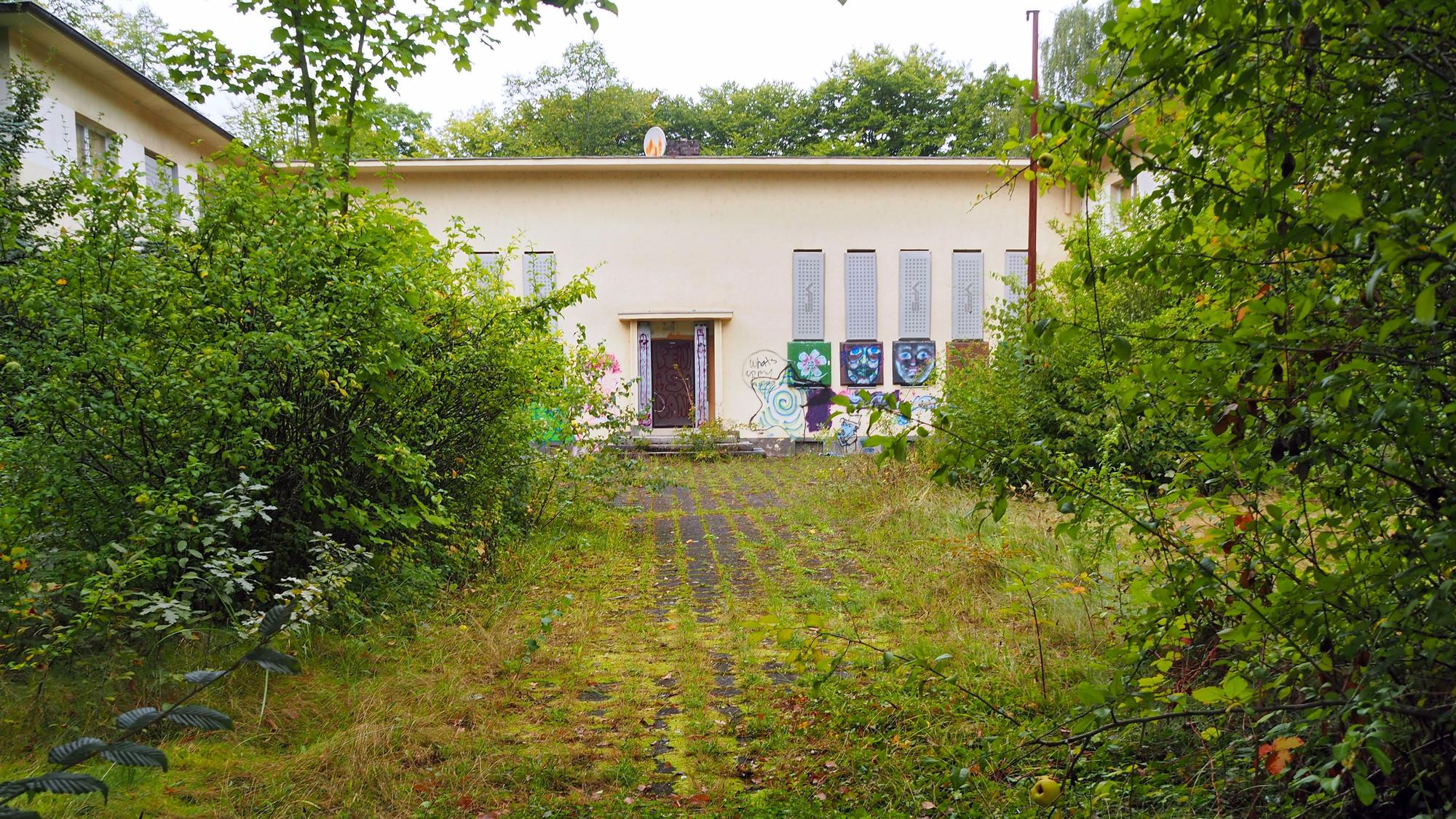 Ein beiges Gebäude mit Flachdach steht hinter Büschen und einer zugewucherten Asphaltfläche.