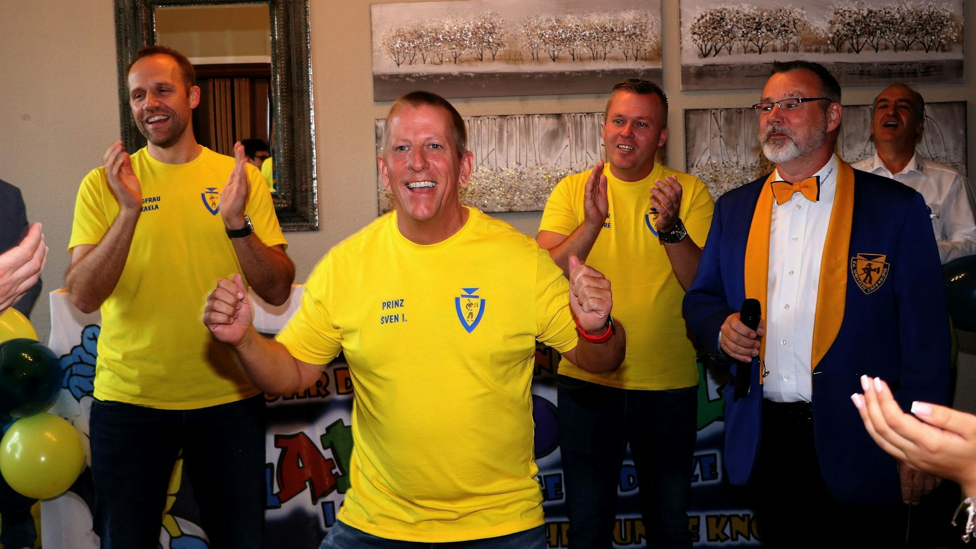 Zu sehen sind drei Herren in gelben T-Shirts und einer mit blau-gelben Anzug und Fliege.