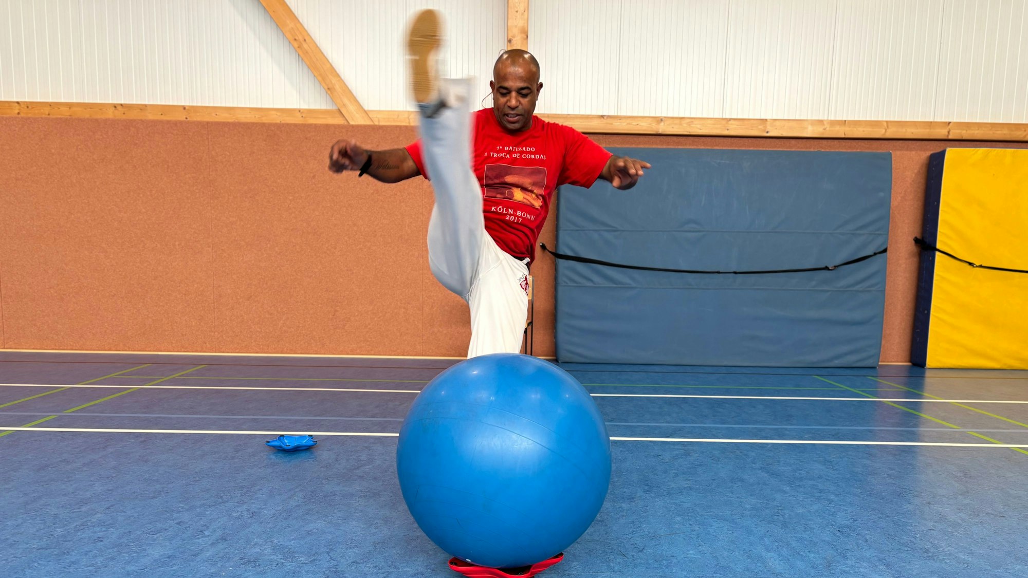 Alexandro Azevedo de Souza tritt im Stand über seine eigene Kopfhöhe. Er befindet sich in einer Turnhalle. Vor ihm ruht ein Gymnastikball auf einem Halter.