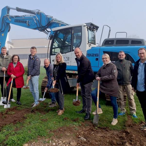 Einige Frauen und Männer stehen vor einem blauen Bagger. Sie haben Schaufeln und Spaten in der Hand, um den symbolischen ersten Spatenstich für die Erweiterung der Grundschule in Blankenheim auszuführen.