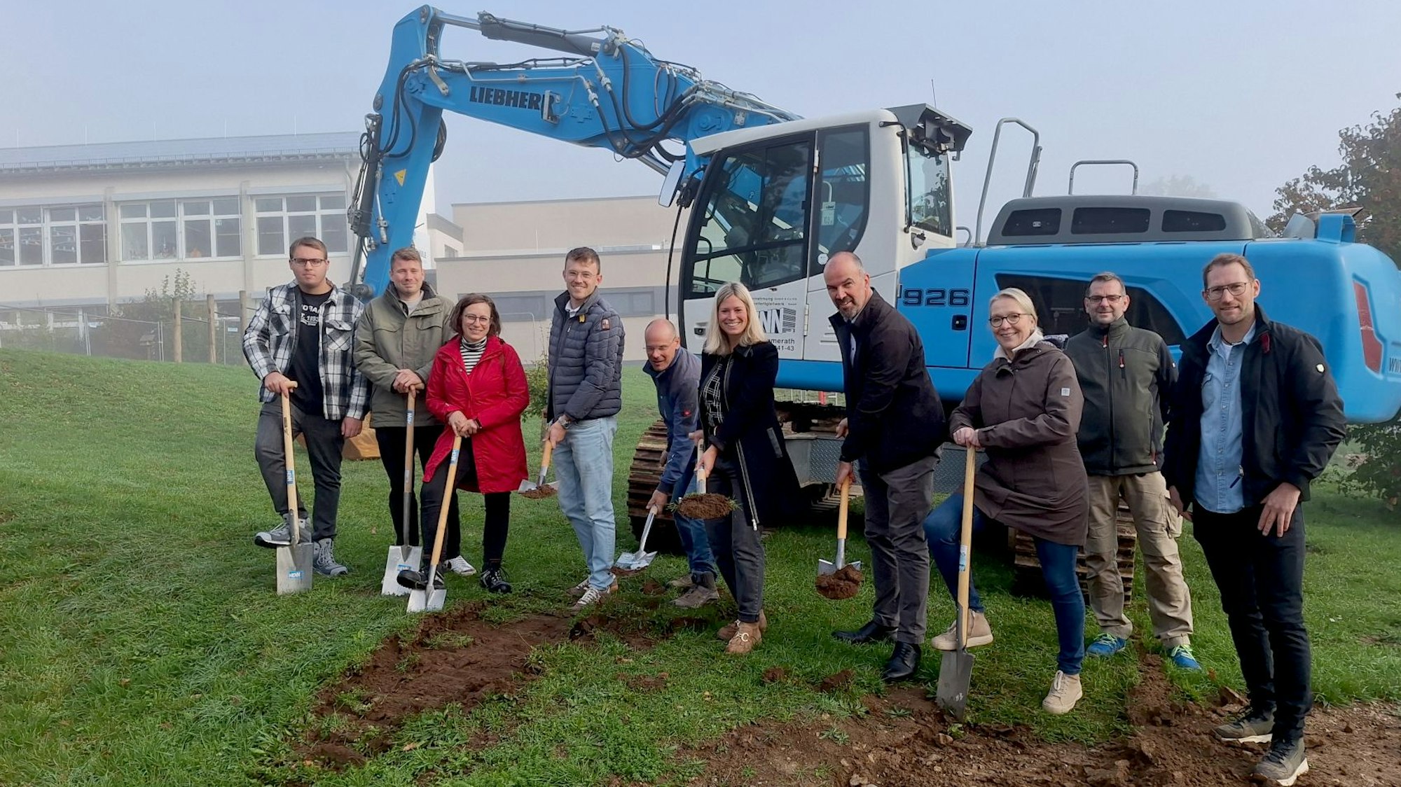 Einige Frauen und Männer stehen vor einem blauen Bagger. Sie haben Schaufeln und Spaten in der Hand, um den symbolischen ersten Spatenstich für die Erweiterung der Grundschule in Blankenheim auszuführen.