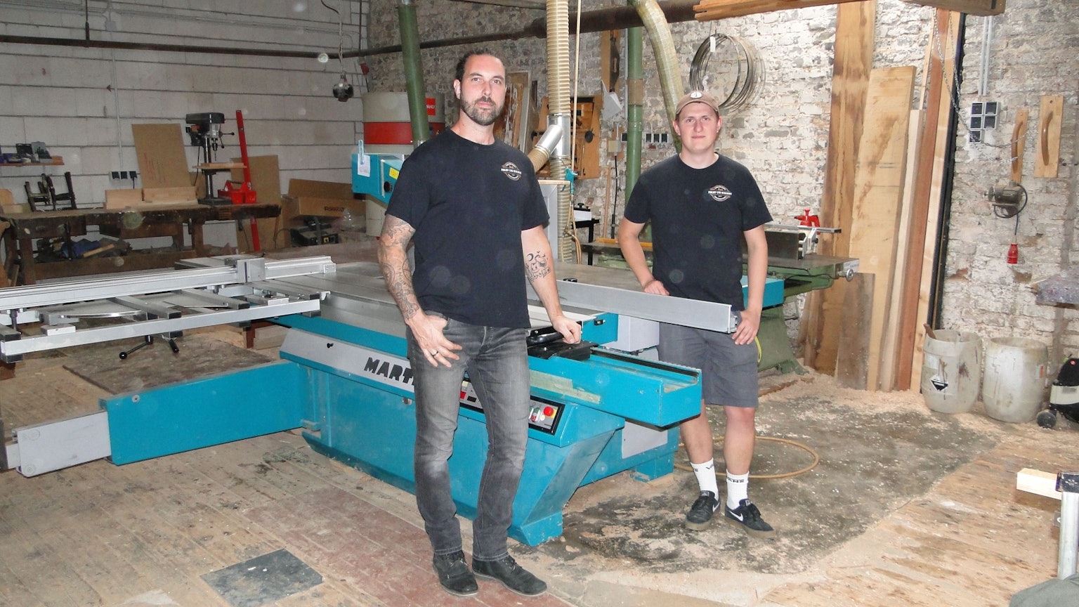 Zwei junge Männer in schwarzen T-Shirts stehen an einer großen Säge in einer Werkstatt.