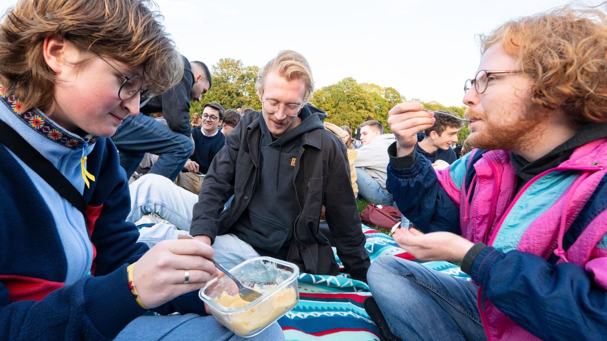 Mehrere Personen sitzen auf einer Picknickdecke und essen ihren Pudding mit einer Gabel.