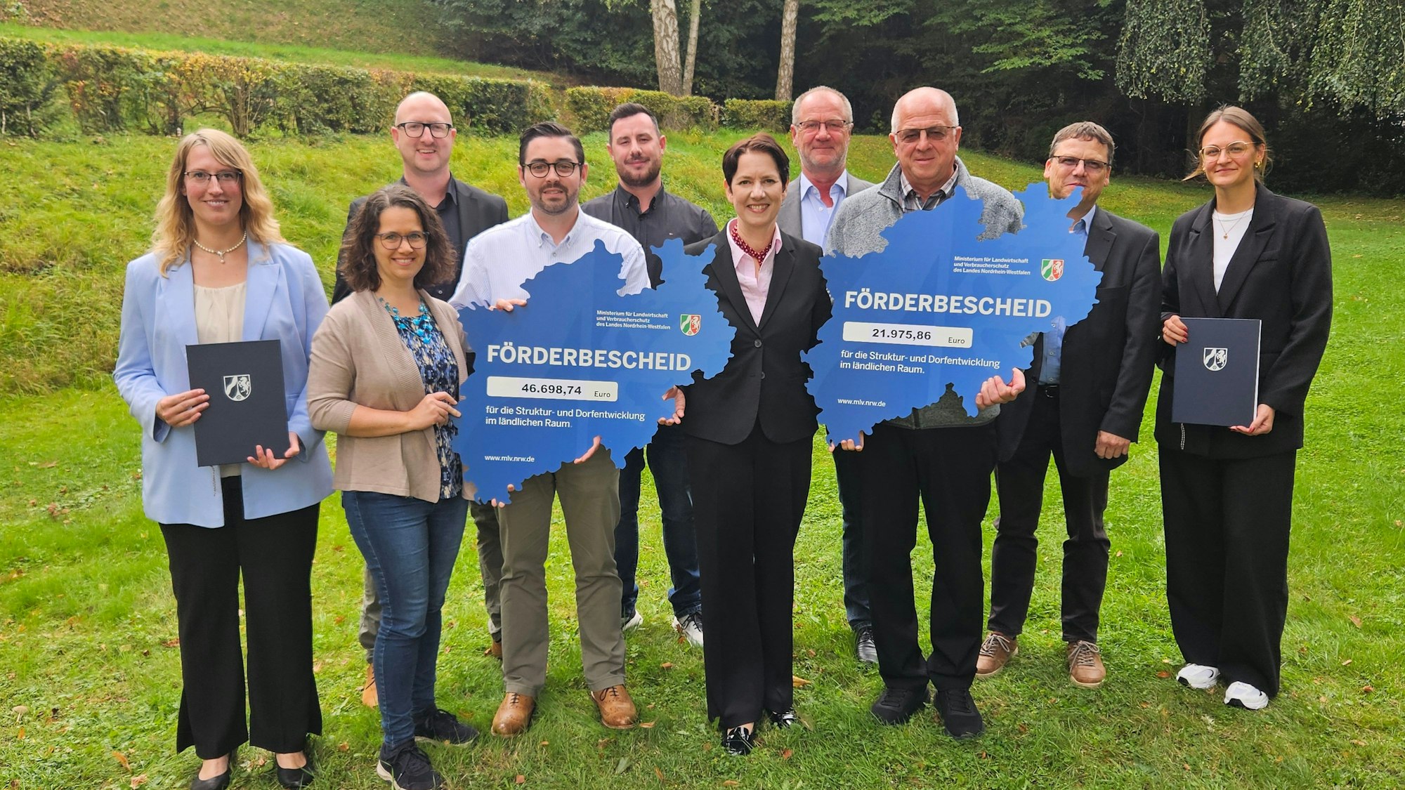 Ministerin Silke Gorißen steht mit Vertretern der Dorfgemeinschaft und des SV Schöneseiffen sowie Bürgermeister Ingo Pfennings und Mitarbeitern der Bezirksregierung Köln nebeneinander und präsentiert zwei symbolische Förderbescheide.