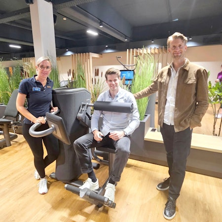 Henning Förster steht neben einem Fitnessgerät, in dem sein Bruder Steffen sitzt. Daneben steht Tanja Hölz.