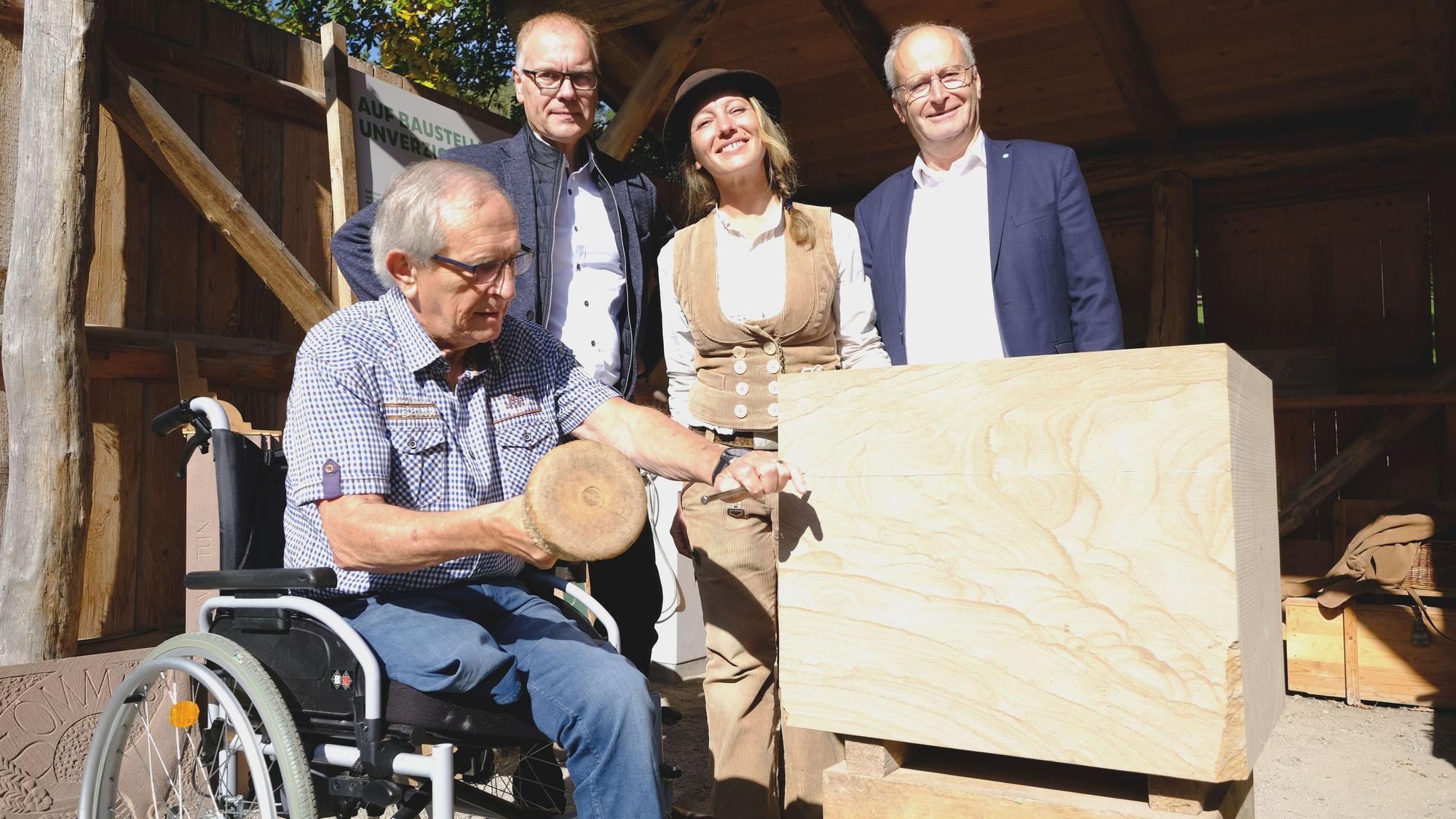 Der im Rollstuhl sitzende Ortsbürgermeister schlägt mit Hammer und Meißel in einen Sandsteinblock. Dahinter stehen der Museumsleiter, die Steinmetzmeisterin und der Bürgermeister.