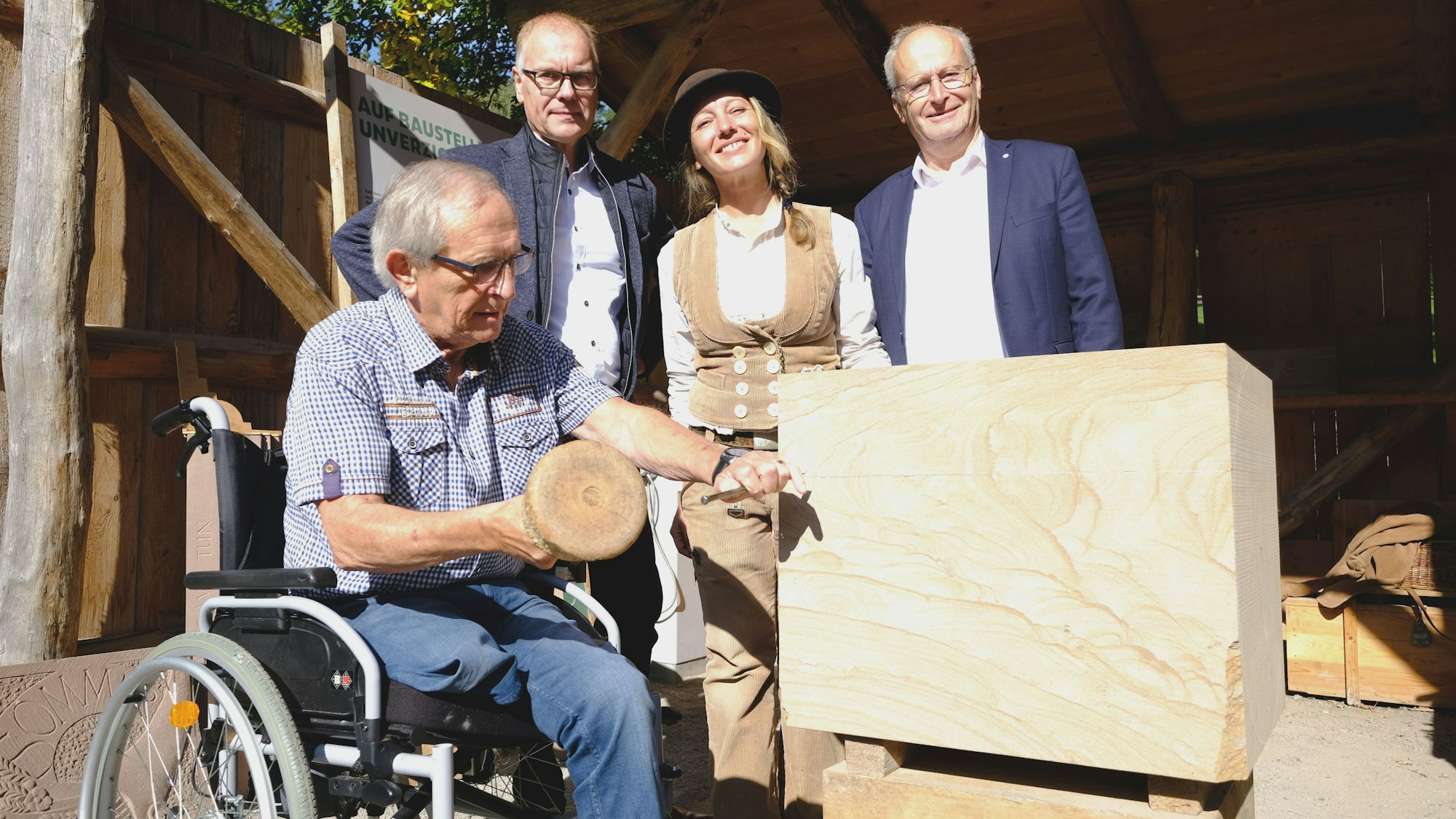 Der im Rollstuhl sitzende Ortsbürgermeister schlägt mit Hammer und Meißel in einen Sandsteinblock. Dahinter stehen der Museumsleiter, die Steinmetzmeisterin und der Bürgermeister.