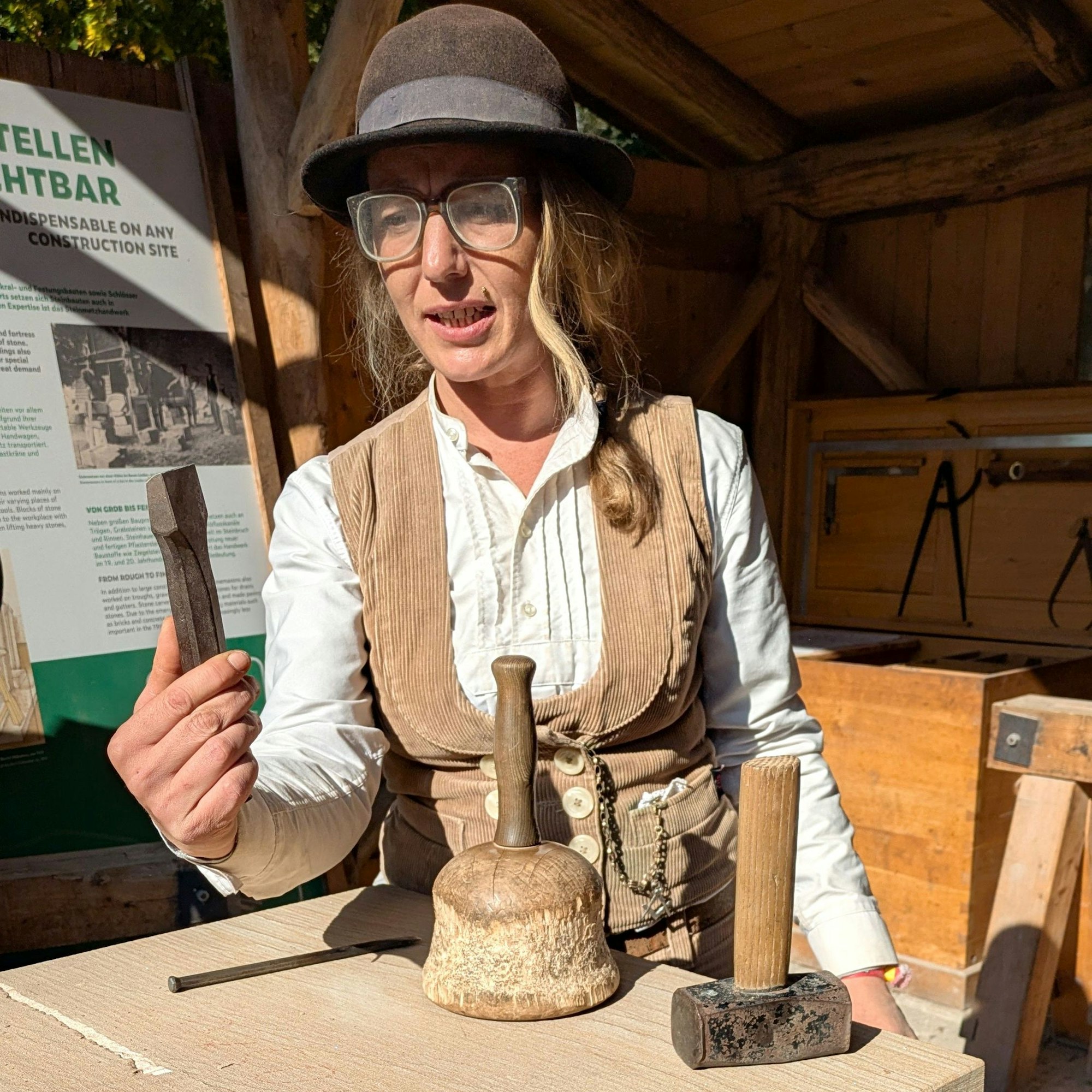 Die Steinmetzmeisterin trägt einen Hut und Arbeitskleidung. Sie hält ein Werkzeug in die Höhe.