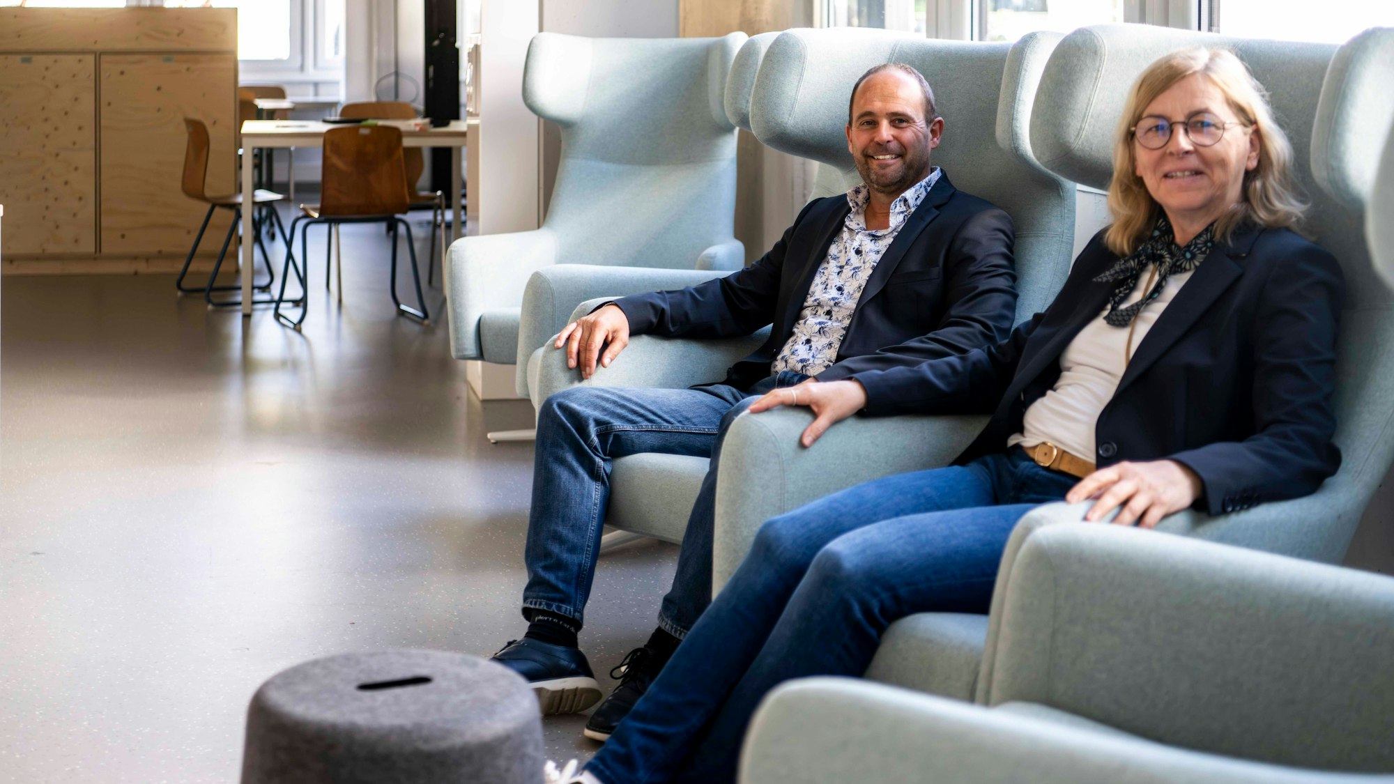 Das Bild zeigt die beiden obengenannten Schulleiter, wie sie in einem grau-grünen Ohrensessel sitzen.