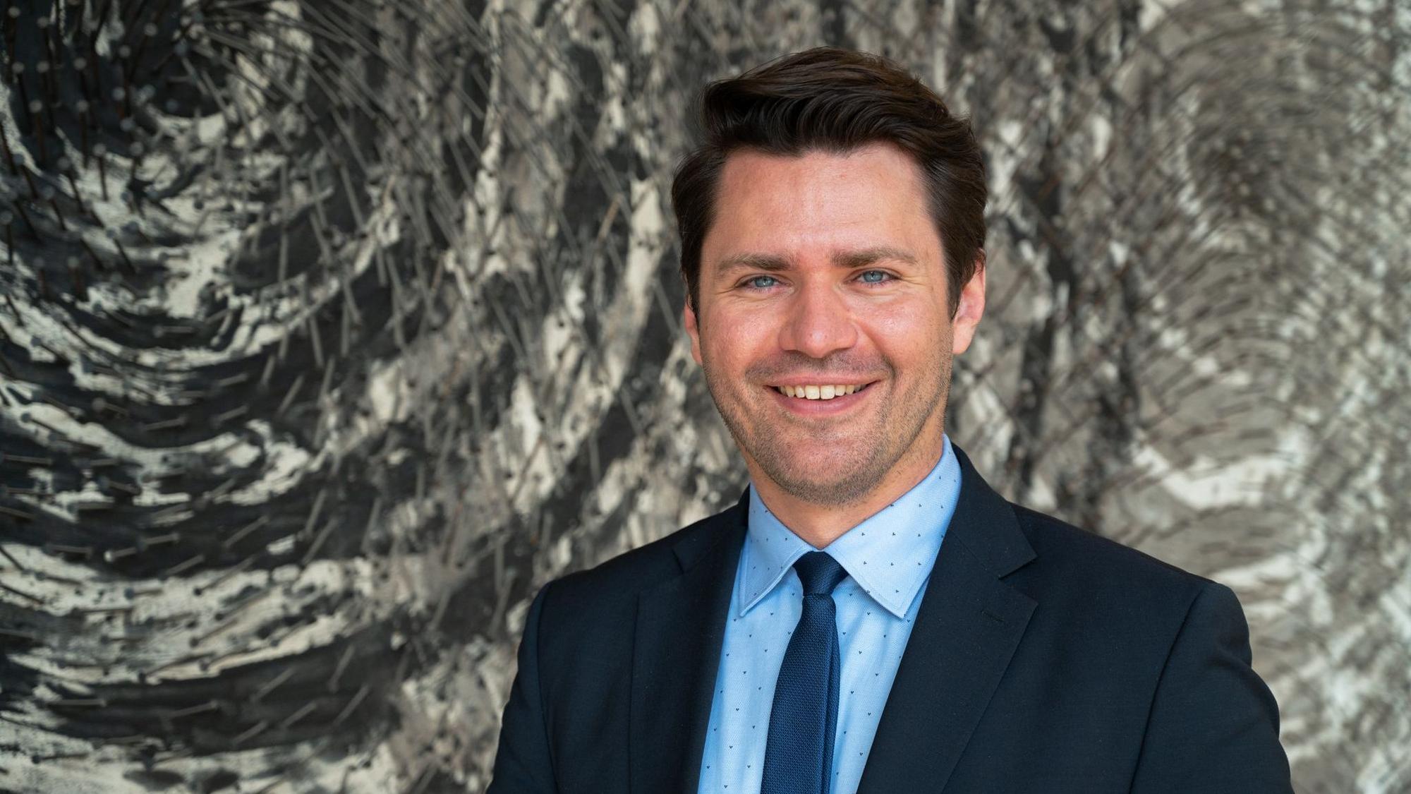 Auf dem Foto ist ein Portrait von Thomas Okos zu sehen. Er trägt einen blauen Anzug.