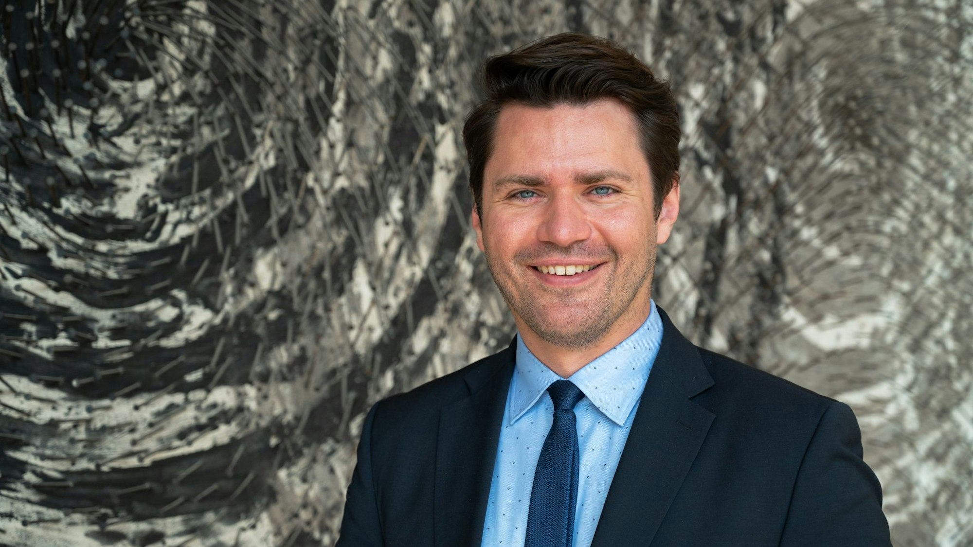 Auf dem Foto ist ein Portrait von Thomas Okos zu sehen. Er trägt einen blauen Anzug.