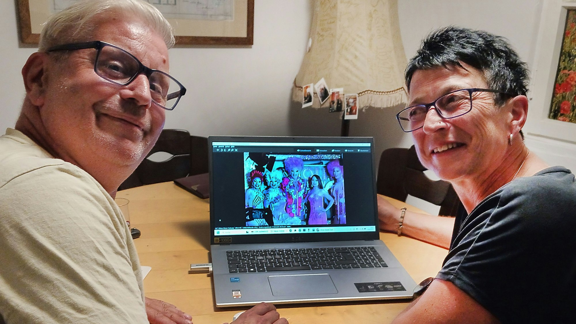 René Berlin und Monika Sauerbier sitzen an einem Tisch, auf dem ein Laptop steht. Auf dem PC ist eine Aufnahme von Dragqueens geöffnet.