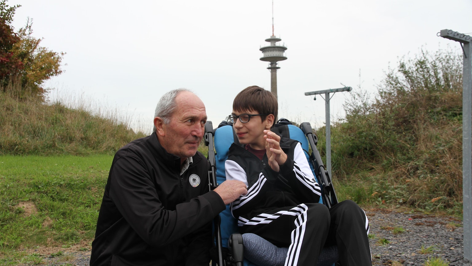 Merab Sharia hockt neben seinem mehrfach behinderten Sohn Bagrat, der in einem Rollstuhl sitzt. Die beiden sind sich vertraut zugewandt.