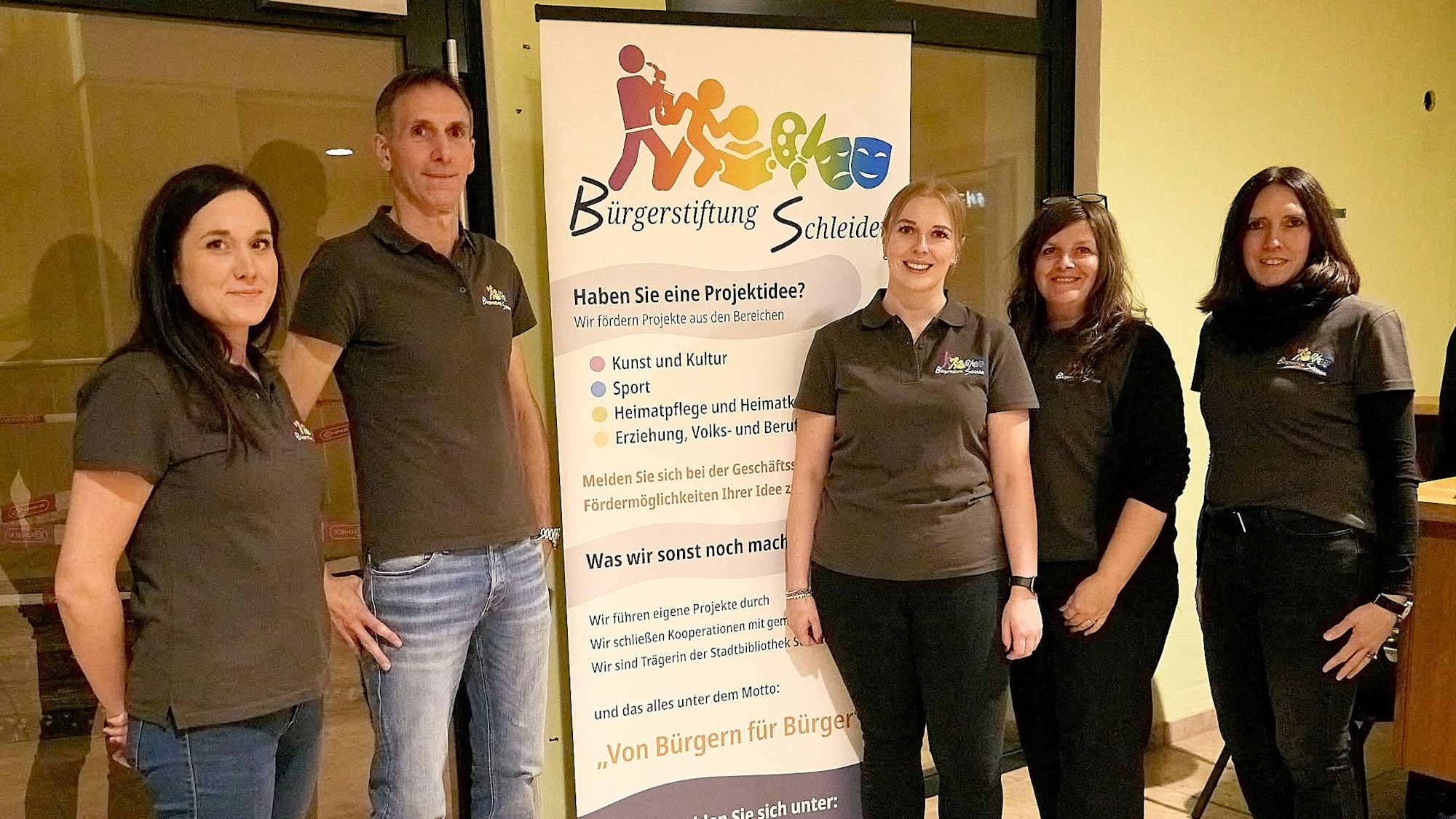 Ein Mann und vier Frauen stehen in identischen Poloshirts neben einem Roll-up-Display der Bürgerstiftung Schleiden.