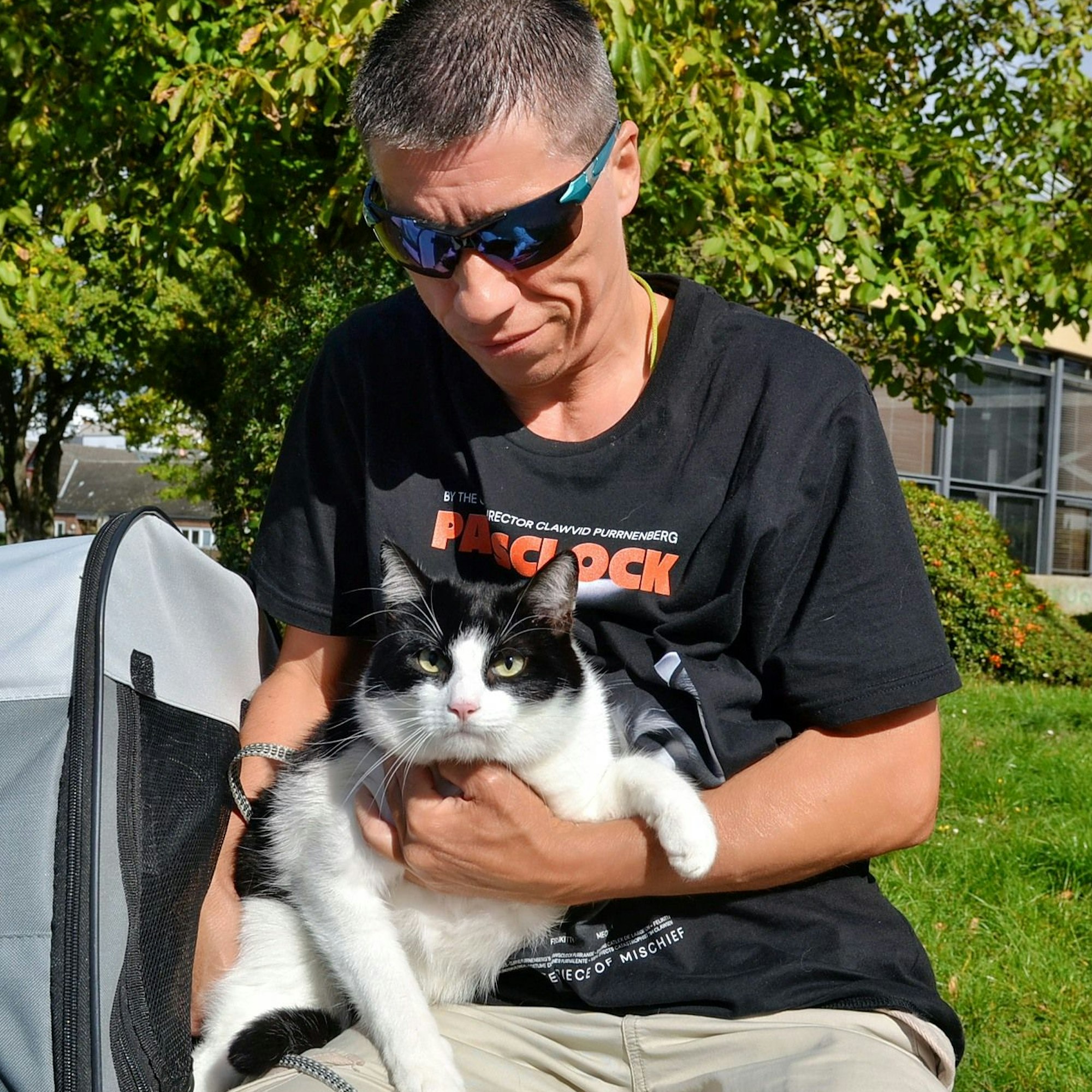 Ein Mann, der ein schwarzes T-Shirt und eine Sportsonnenbrille trägt, hält einen schwarz-weißen Kater auf dem Arm.
