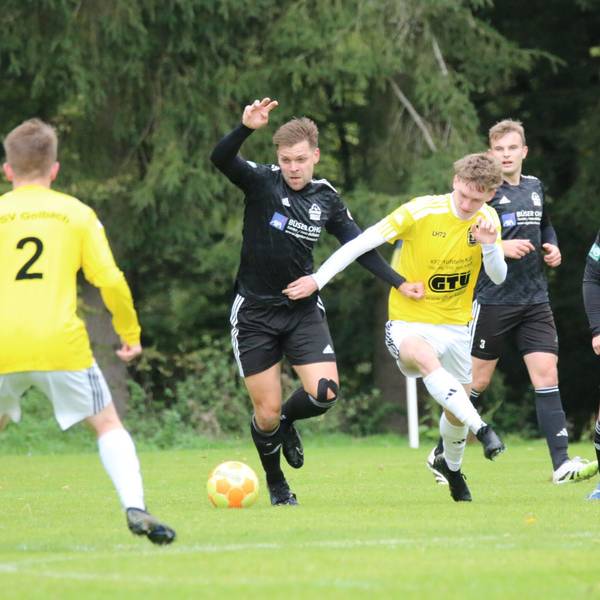 Zwei Fußballer kämpfen um den Ball, mehrere Mitspieler beobachten das Duell.