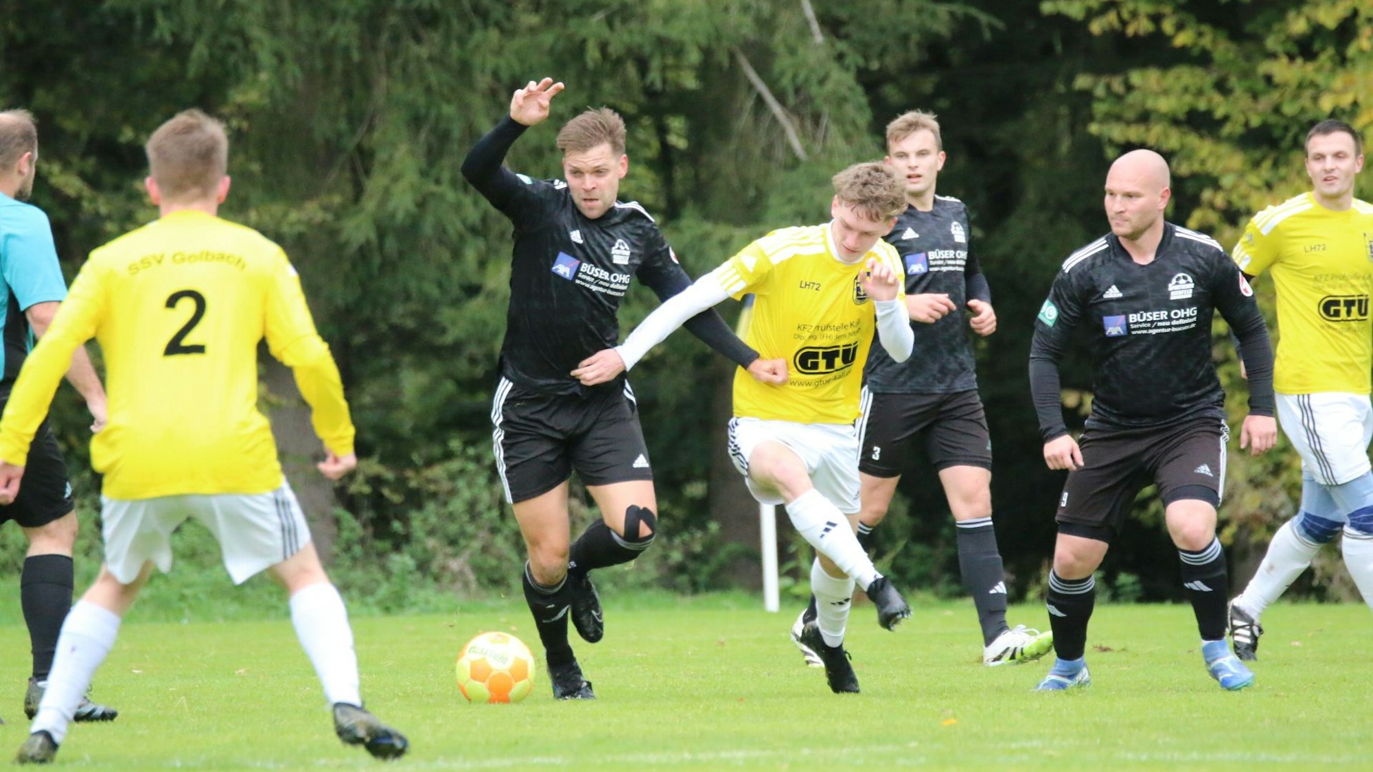 Zwei Fußballer kämpfen um den Ball, mehrere Mitspieler beobachten das Duell.