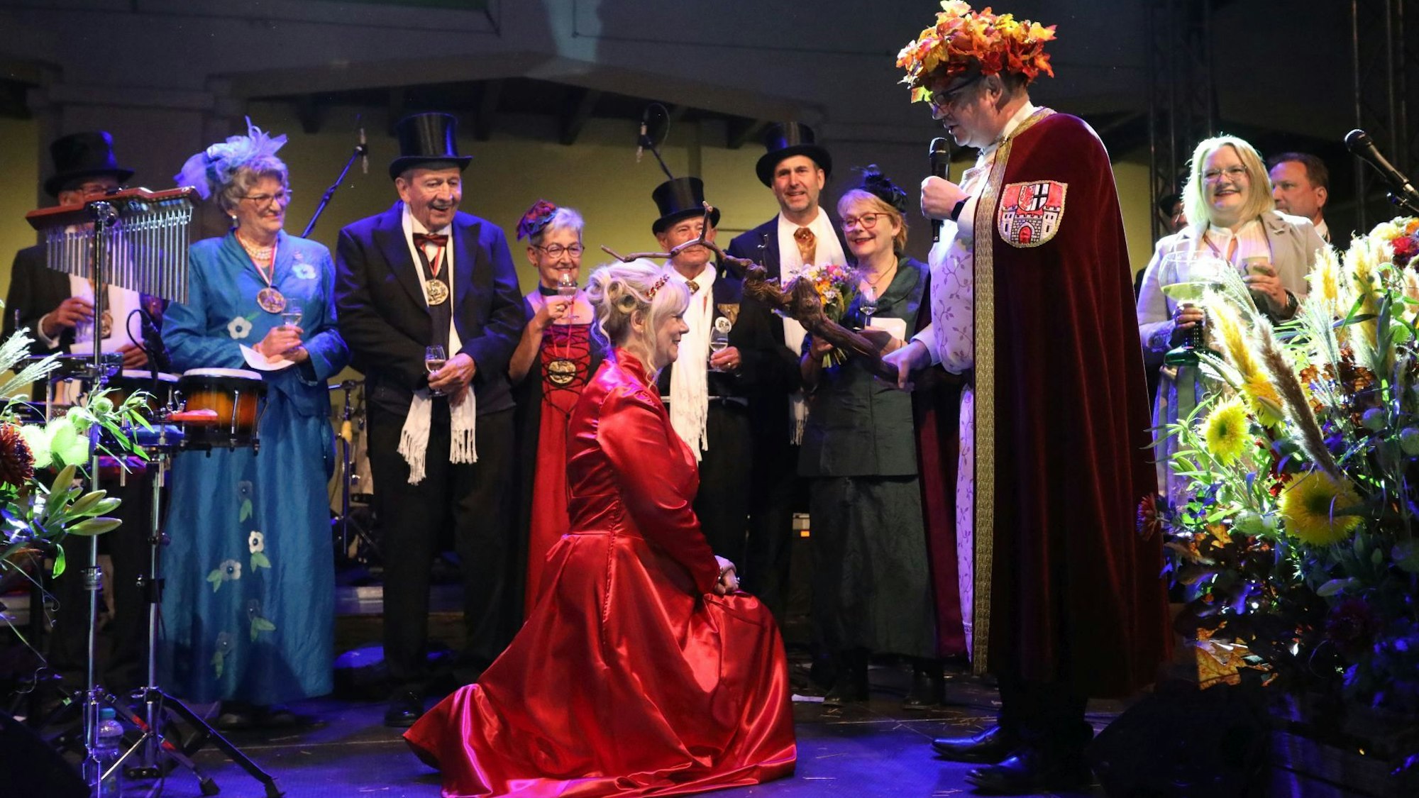 Eine Frau in rotem Kleid kniet vor einem Mann, der eine „Krone“ aus Weinlaub trägt.