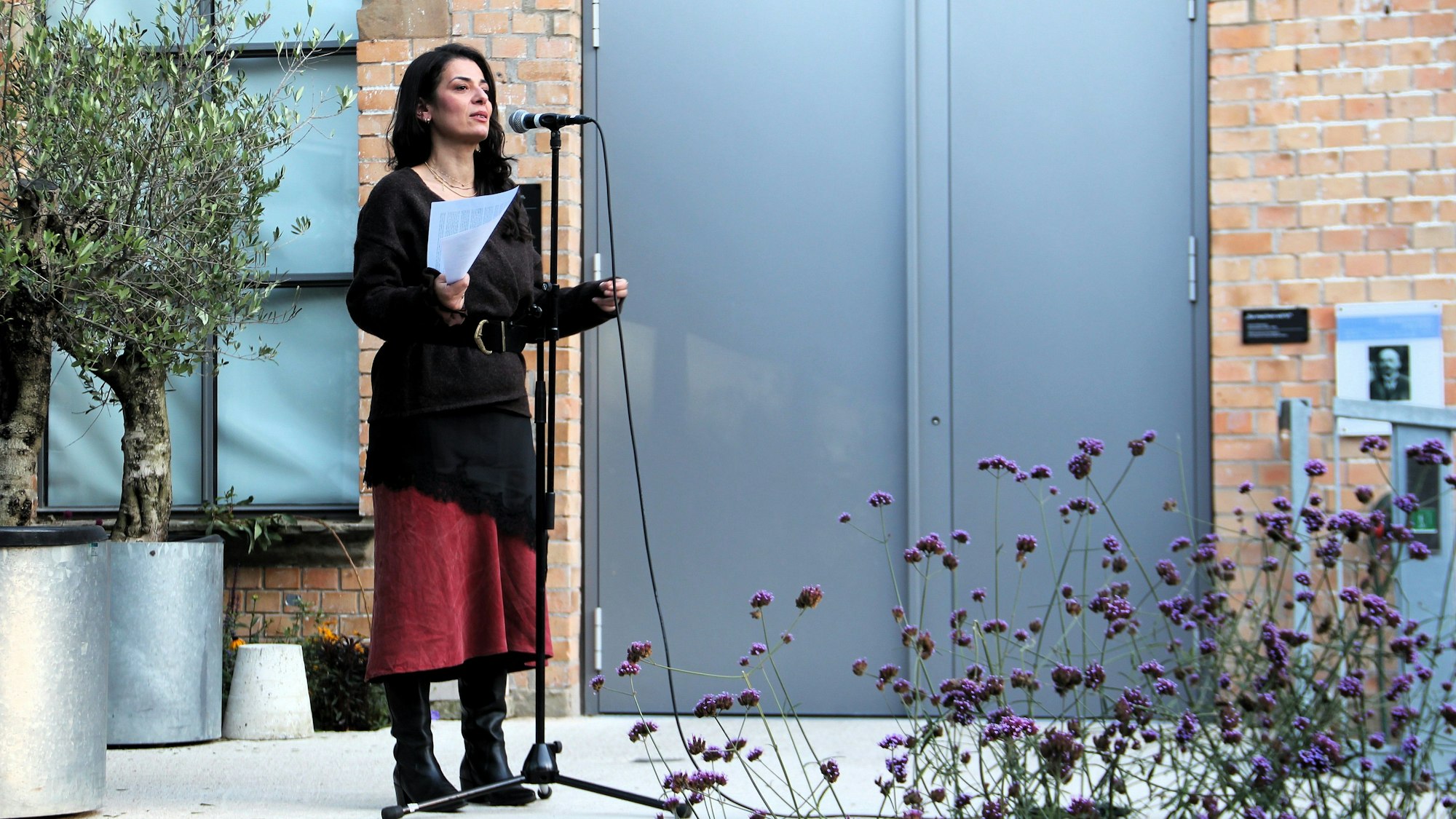 Eine Frau mit dunklem Haar steht vor einem Mikrofon, sie hält einen Zettel in der Hand.