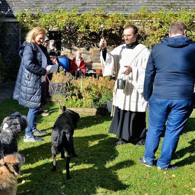 Diakon Francesco Tabacco, der ein liturgisches Gewand trägt, besprengt mehrere Hunde, die von ihren Frauchen an der Leine gehalten werden, mit Weihwasser.