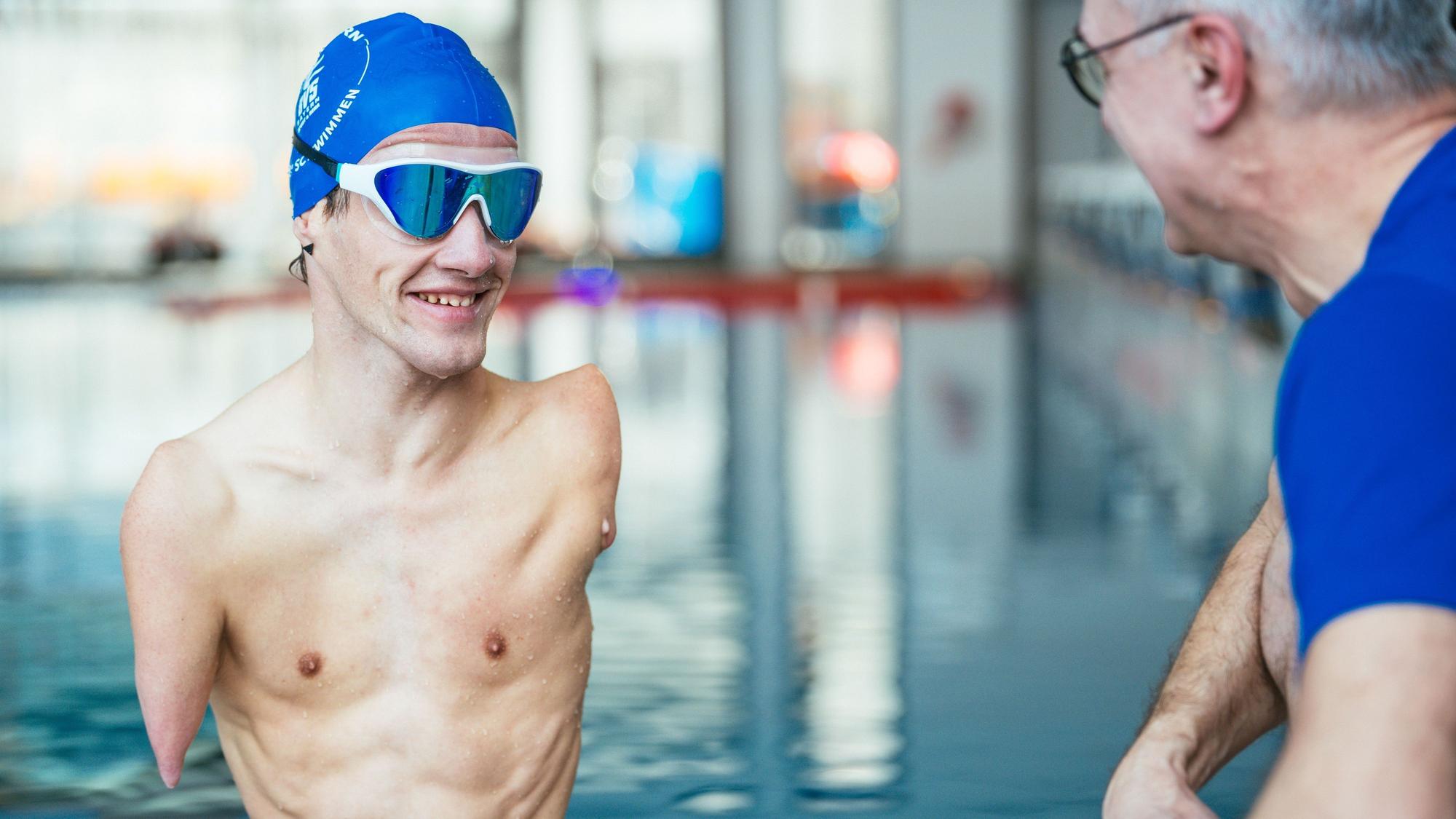 Janis McDavid trägt eine blaue Haube und eine Schwimmbrille. Neben ihm ein Mann mit blauem T-Shirt. Im Hintergrund ein Schwimmbad.