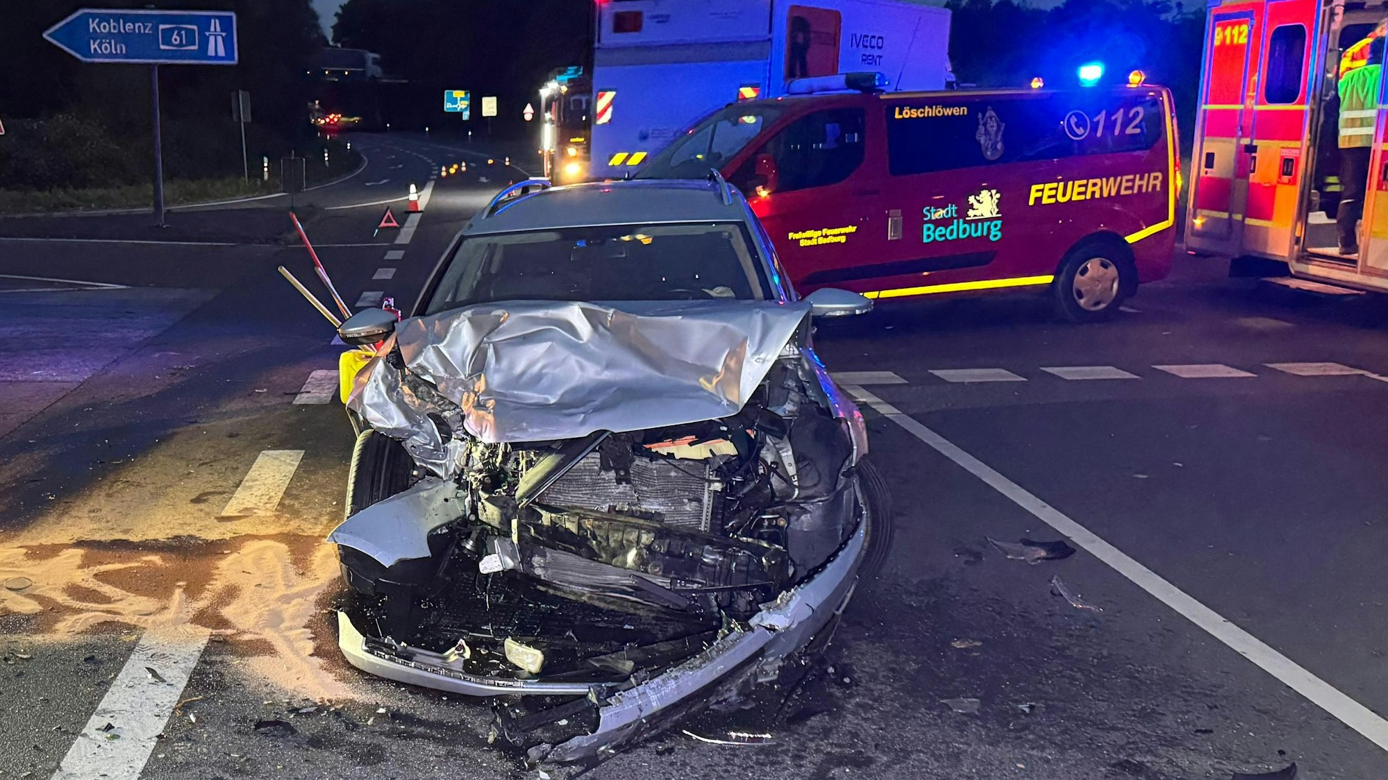 Ein Auto mit zerstörtem Motorraum und Karosserie. Im Hintergrund Feuerwehr und Rettungswagen.