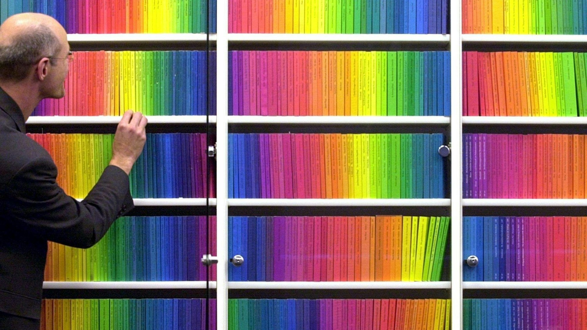 Ein Mann greift nach einem der vielen Taschenbücher in Regenbogenfarben, die mehrere Regalmeter ausmachen.