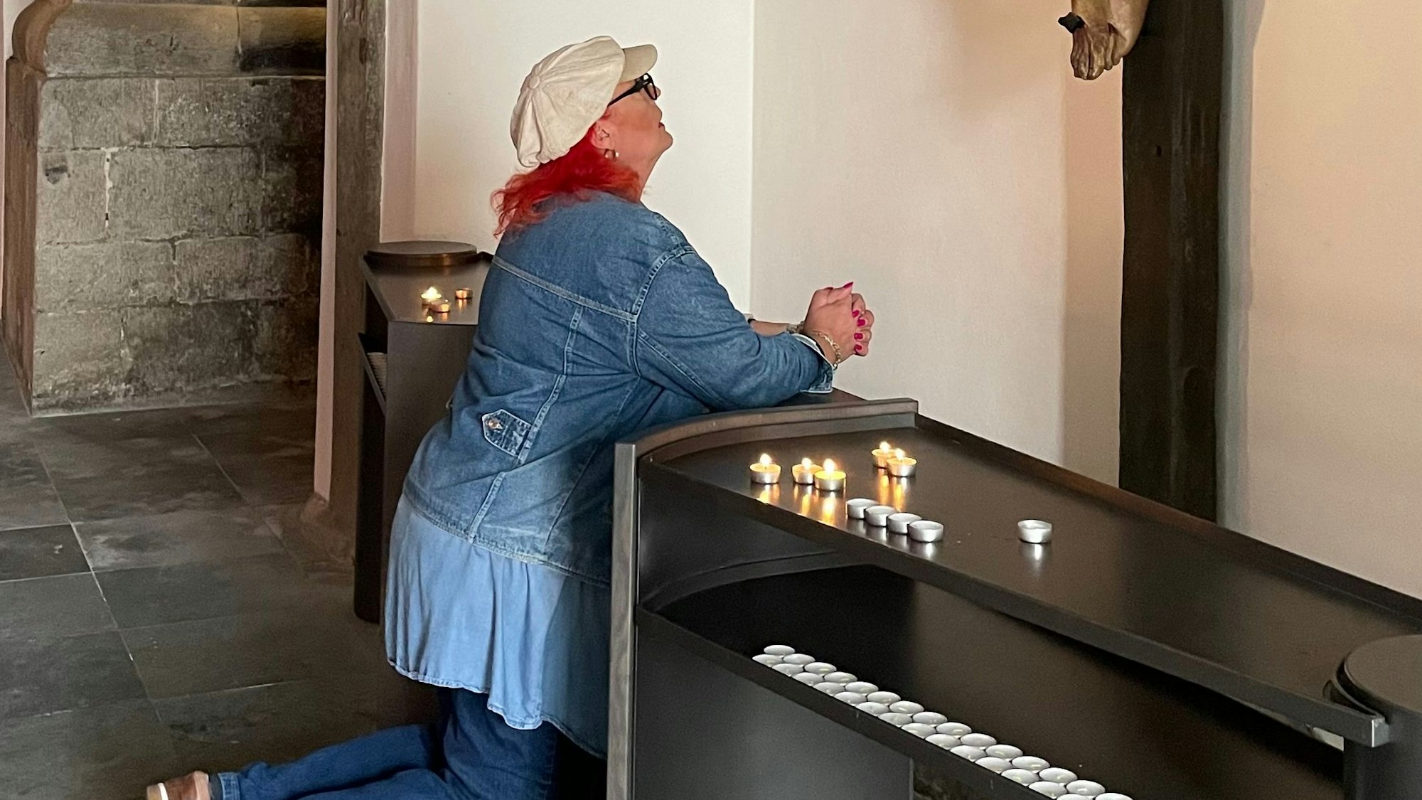 Vor einem Kreuz kniet eine Frau mit rotem Haar mit gefalteten Händen.
