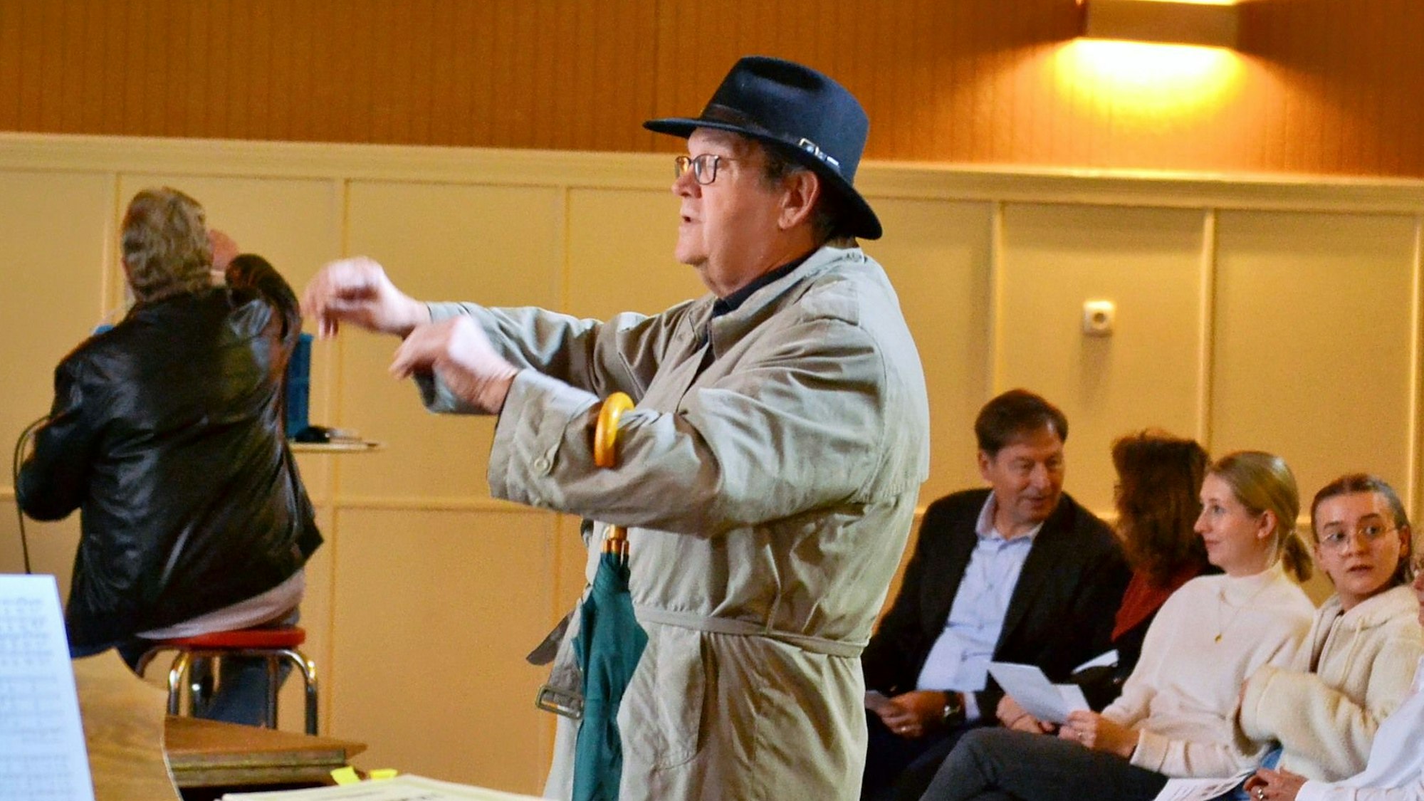 Der Dirigent trägt einen Mantel und einen Hut, über dem Arm hängt ein Regenschirm.