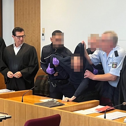 Drei Justizwachtmeister zwingen den Angeklagten, aufzustehen. Er hatte sich geweigert, sich beim Eintreten der Kammer in den Saal zu erheben und das Gericht beleidigt. Links Verteidiger Michael Hafner, rechts Verteidiger Bernd Arnold.