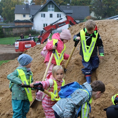 Mehrere Kinder mit Schaufeln auf einem Sandhügel, im Hintergrund ist ein Bagger zu sehen.