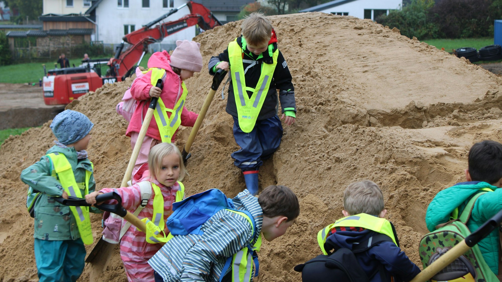 Mehrere Kinder mit Schaufeln auf einem Sandhügel, im Hintergrund ist ein Bagger zu sehen.