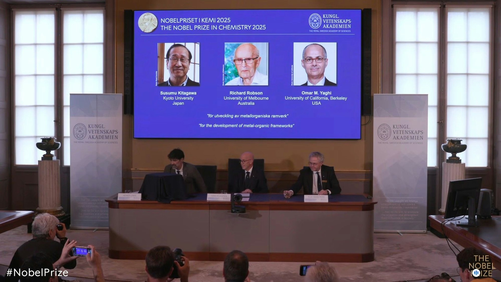 Standbild aus dem Livestream zur Bekanntgabe des Nobelpreises für Chemie 2025: Auf einer Leinwand ist die Präsentation der diesjährigen Preisträger, Susumu Kitagawa (Japan), Richard Robson (Australien) und Omar Yaghi (USA), zu sehen.