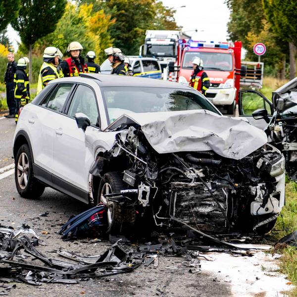 Nach einem Verkehrsunfall stehen zwei stark demolierte Autos auf der Straße, im Hintergrund sind Einsatzkräfte der Feuerwehr zu erkennen.