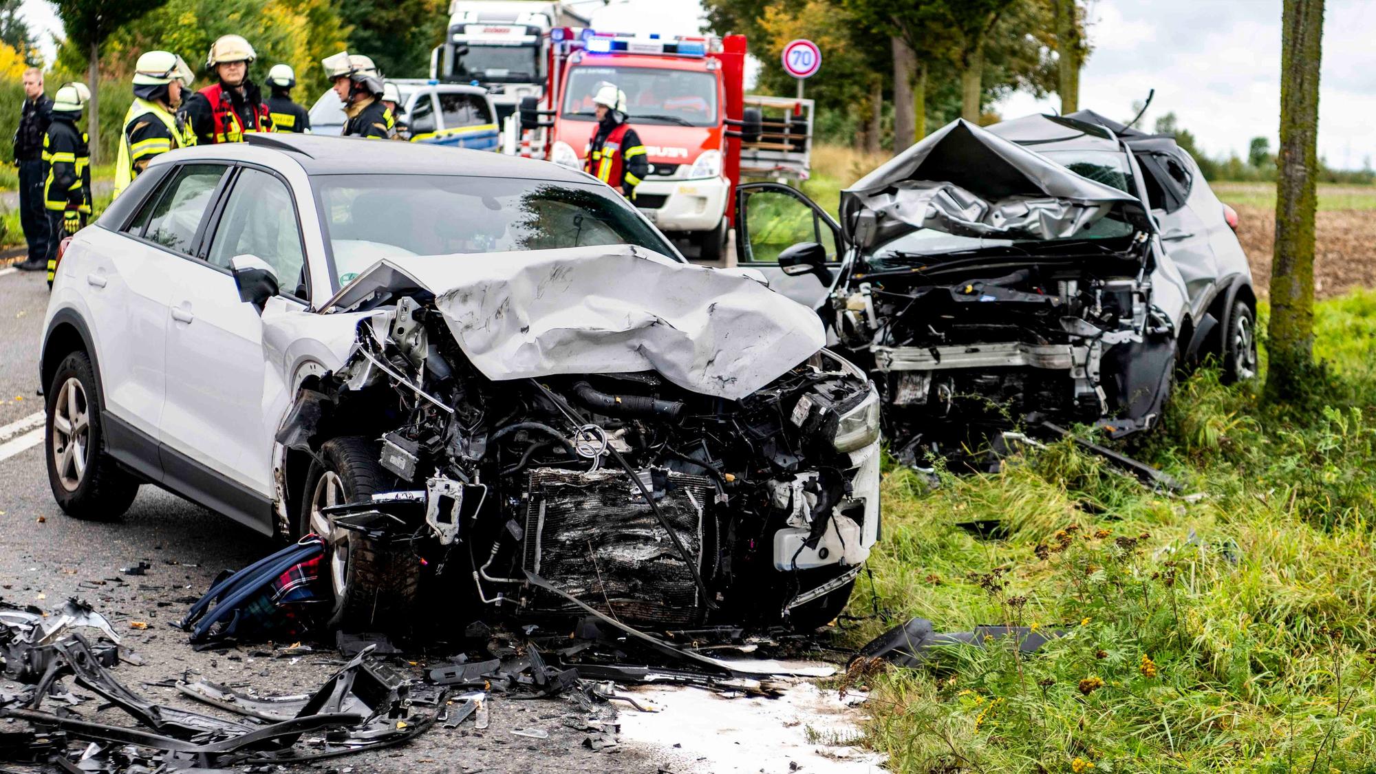Nach einem Verkehrsunfall stehen zwei stark demolierte Autos auf der Straße, im Hintergrund sind Einsatzkräfte der Feuerwehr zu erkennen.
