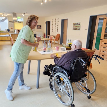 Eine Pflegekraft reicht einem im Rollstuhl sitzenden Mann ein Glas zum Trinken.