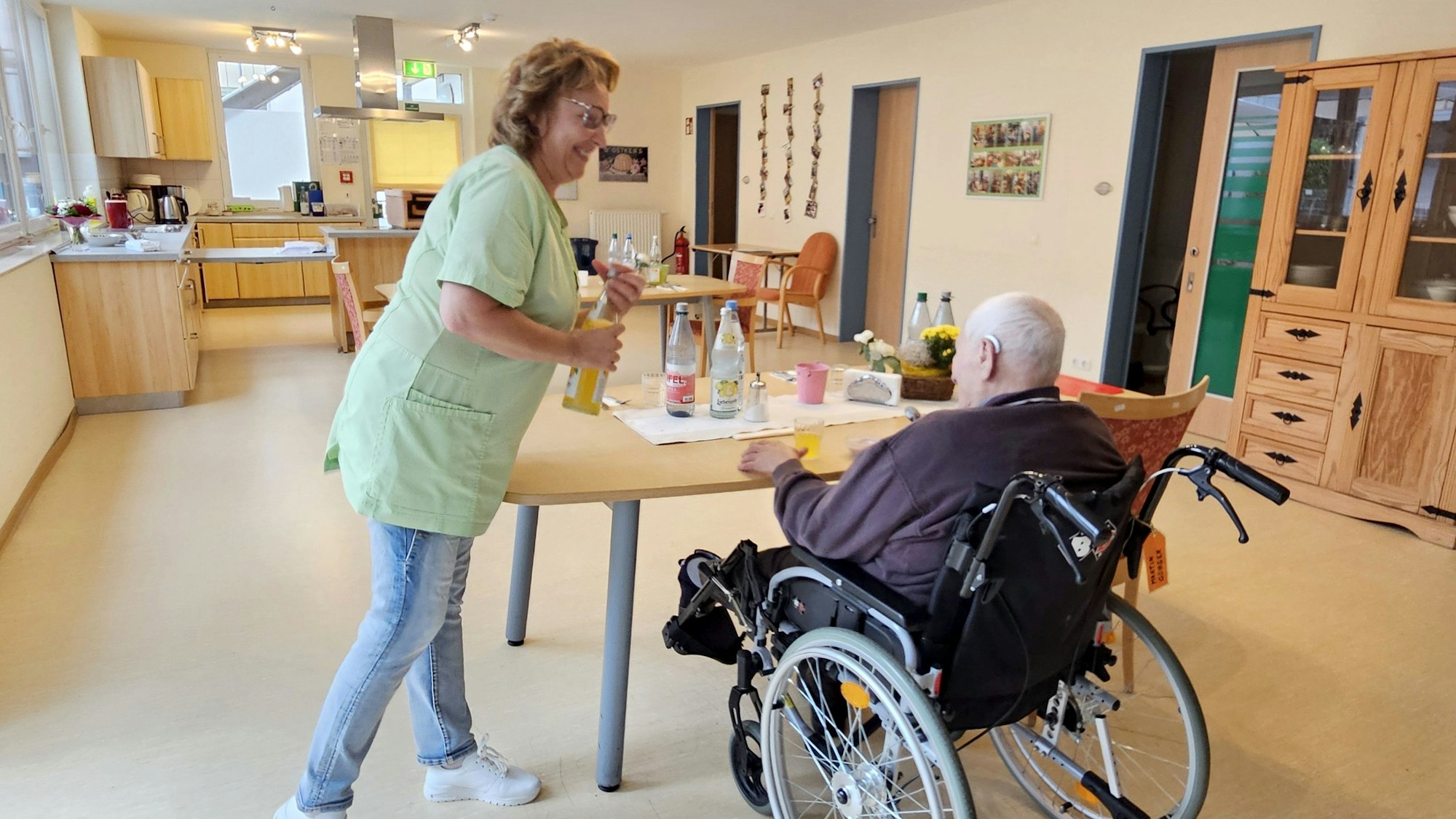 Eine Pflegekraft reicht einem im Rollstuhl sitzenden Mann ein Glas zum Trinken.