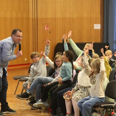 Ein Mann mit Mikrofon vor Schülerinnen und Schülern, die sich melden.