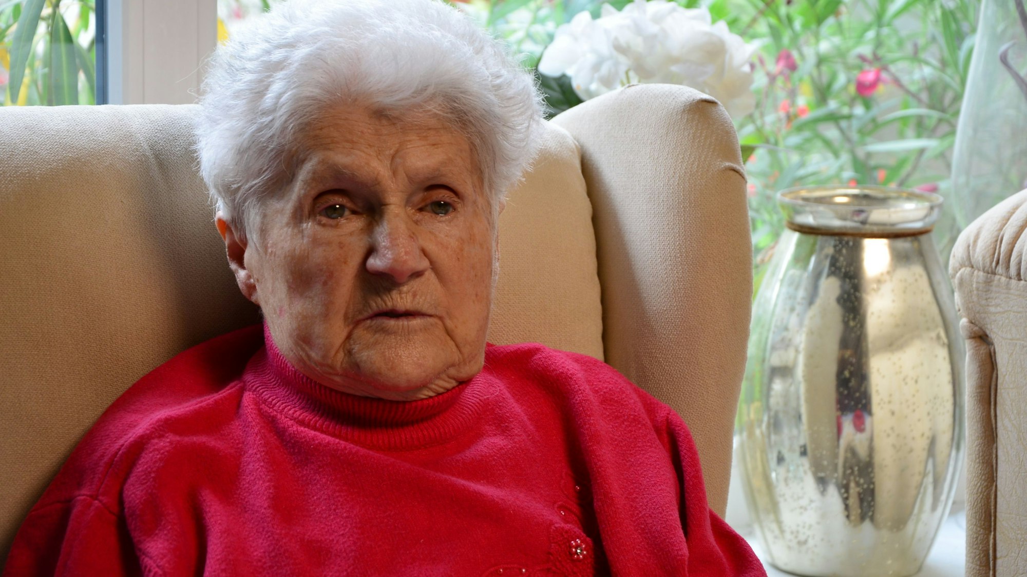 Das Bild zeigt die Dame auf ihrem Sessel. Sie trägt einen roten Pullover.