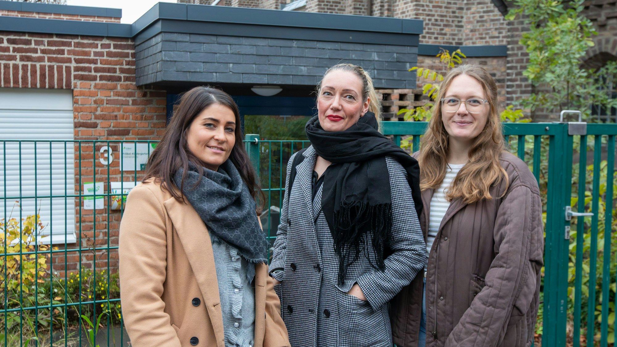 Drei Frauen stehen vor einem grünen Zaun, hinter dem eine Kita liegt.