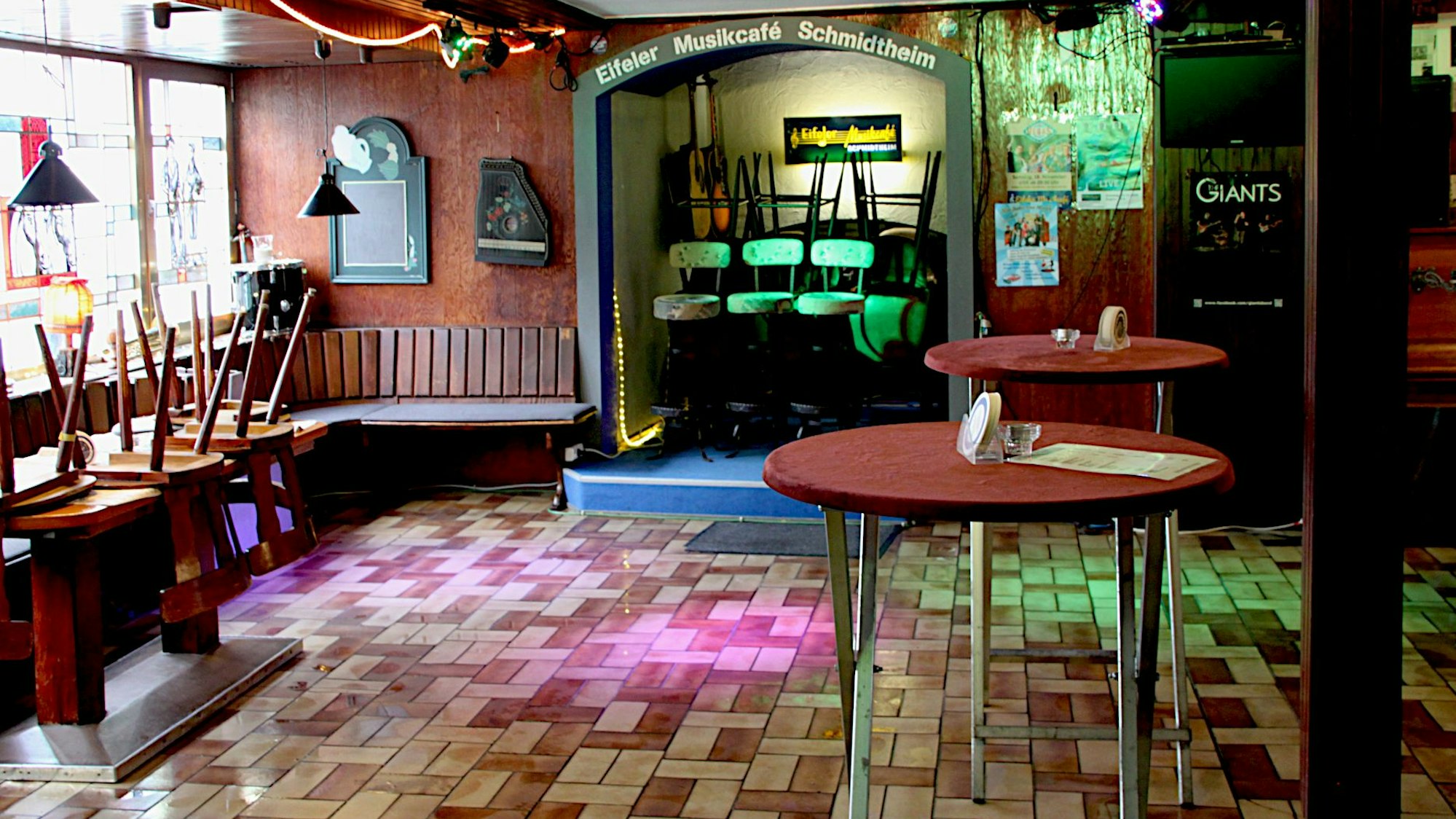 Blick in das „Eifeler Musikcafé“: Der Boden in der ehemaligen Gaststätte ist gefliest. Buntes Licht aus Scheinwerfern leuchtet darauf. Im Raum befinden sich zwei Stehtische.