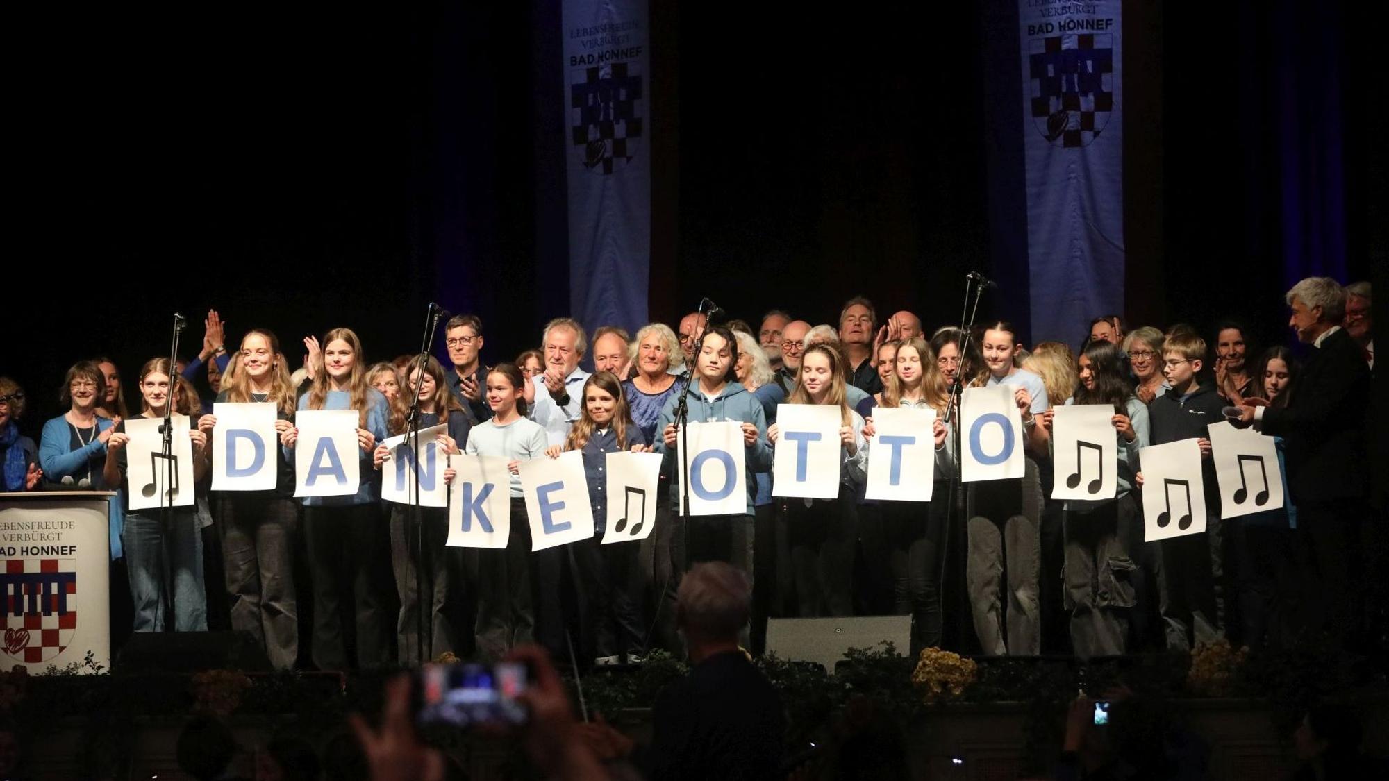 Zwei Chöre auf der Bühne, die in der ersten Reihe auf großen Blättern mit einzelenen Buchstaben „Danke Otto“ zeigen.