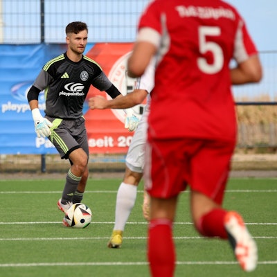 Torwart Jan Beyers hat den Ball am Fuß und schaut nach vorne, um sich einen Überblick zu verschaffen. Im Vordergrund ist unscharf ein Verteidiger von hinten zu sehen.