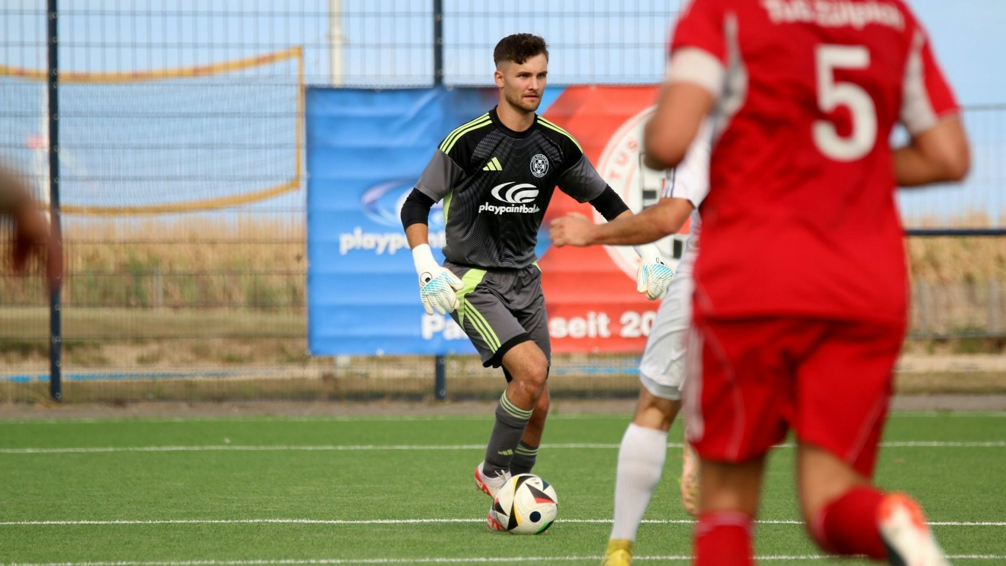 Torwart Jan Beyers hat den Ball am Fuß und schaut nach vorne, um sich einen Überblick zu verschaffen. Im Vordergrund ist unscharf ein Verteidiger von hinten zu sehen.