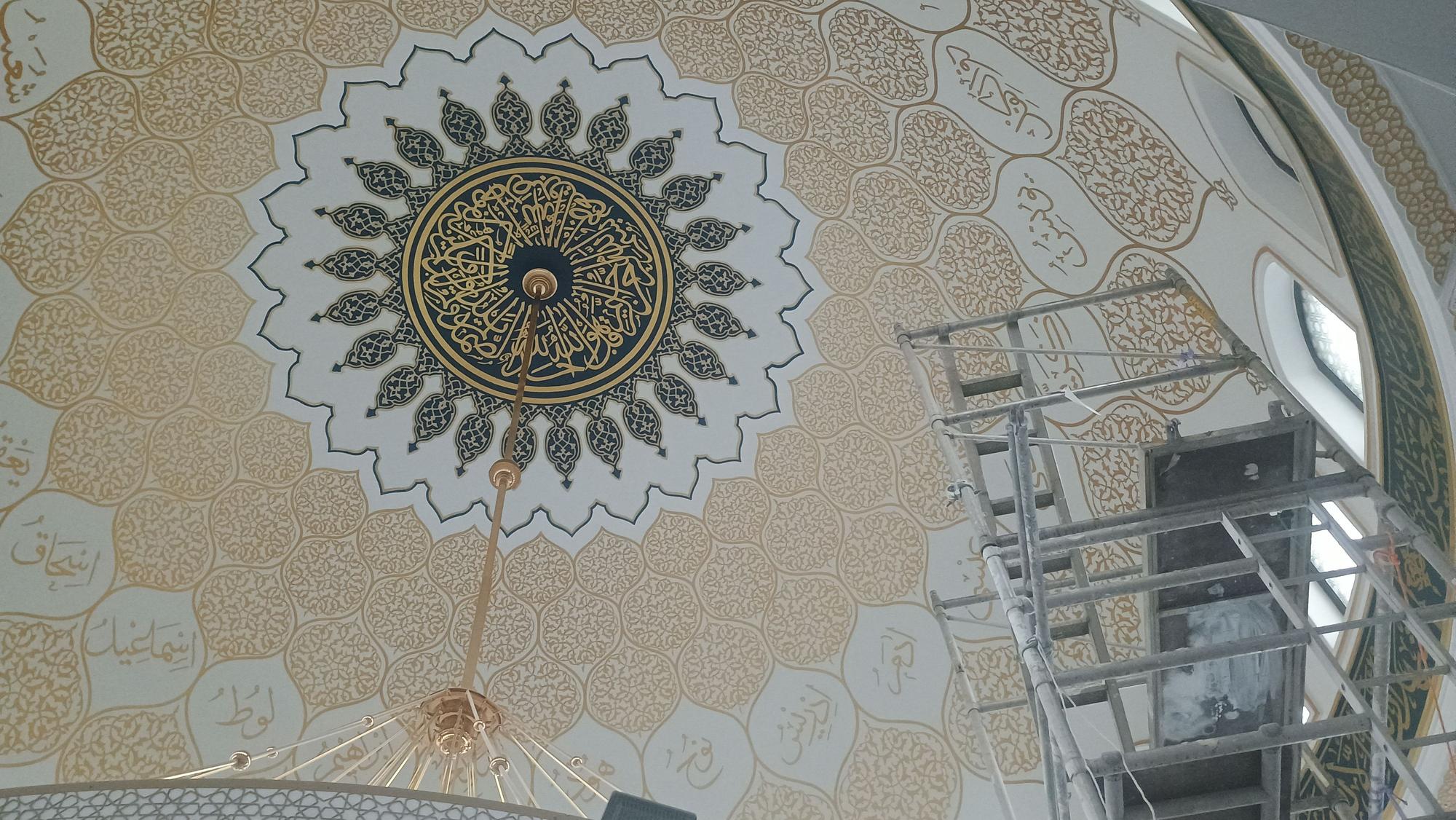 Wer nach oben in die Mitte des Kuppeldachs blickt, sieht ein kreisförmiges Medaillon, ebenfalls mit goldener Kalligrafie geschmückt, und drumherum ein florales Muster, das sternförmig über die gesamte Kuppel verläuft.
