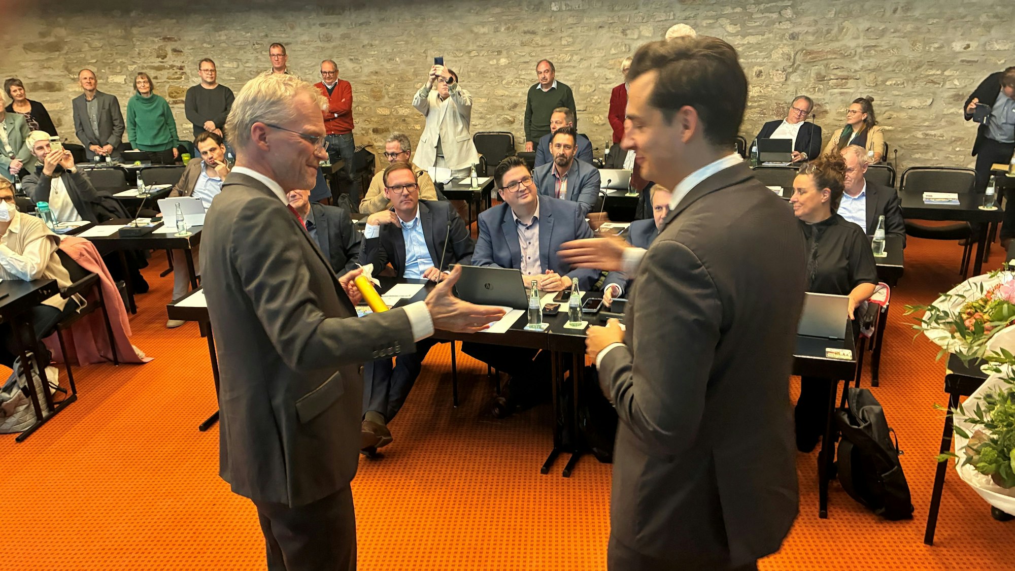 Zwei Männer im Anzug stehen in einem Ratssaal. Der eine überreicht dem anderen einen Staffelstab.