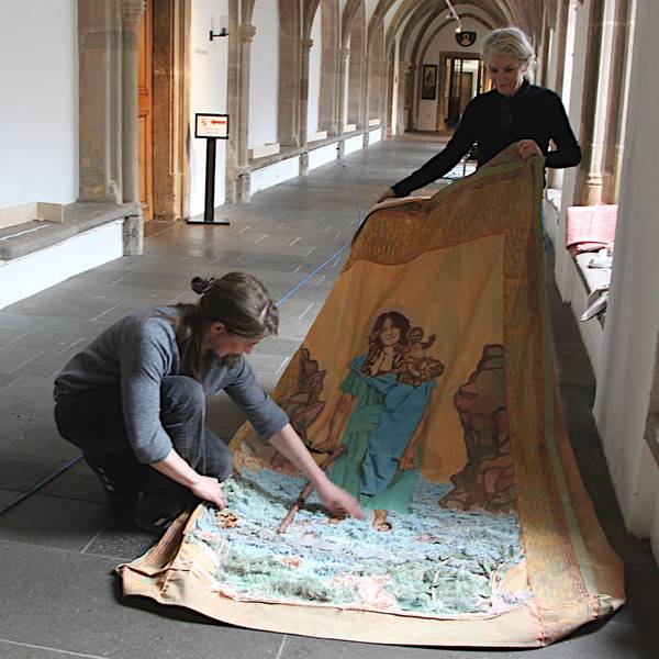 Monika von Bernuth (l.) und Vera Sous halten ein großes Stück Stoff, auf dem Ghada Khano als heiliger Christopherus dargestellt wird. Sie befinden sich im Kreuzgang von Kloster Steinfeld.